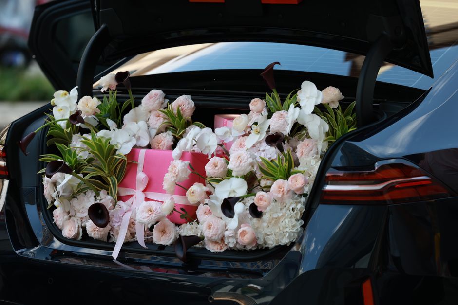 A car trunk filled with a lush floral arrangement consisting of white and pale pink roses, white orchids, dark purple calla lilies, and green foliage, with two pink gift boxes tied with white ribbons