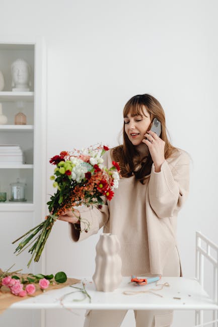 A woman with light brown hair and bangs, dressed in a beige sweater, stands indoors at a white table, holding a vibrant bouquet of fresh flowers including white, red, pink, and green blooms. She is sm