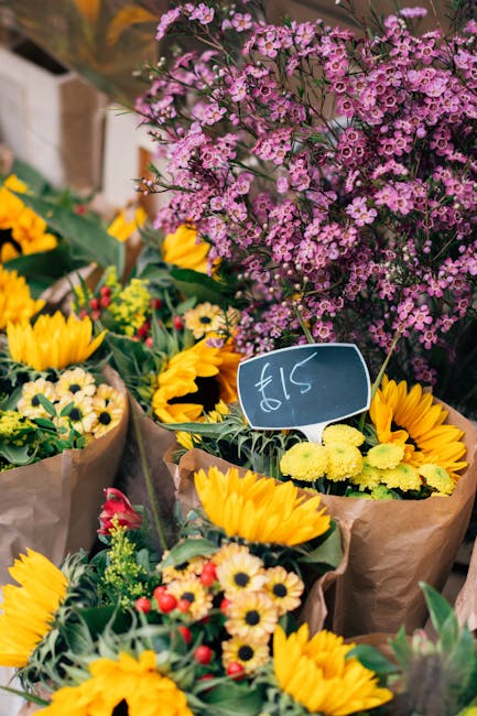 A vibrant display of fresh flowers at a local flower market, featuring bright yellow sunflowers with large, round petals and dark centers, surrounded by smaller cream and red daisies. Behind them, a b