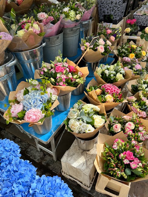 A variety of fresh floral bouquets arranged in metal buckets and wrapped in brown craft paper, featuring flowers such as pink roses, white carnations, pale blue hydrangeas, and other colorful blooms.