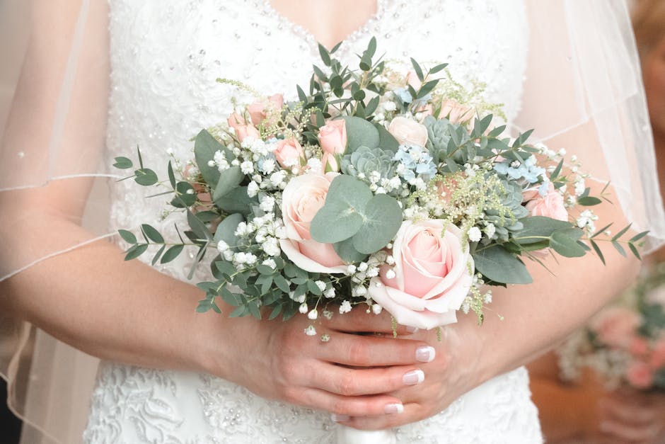 A bride holds a floral bouquet featuring pale pink roses, white baby's breath, and silver-green eucalyptus leaves, arranged in a rounded, lush style. She is wearing a white wedding dress with intricat
