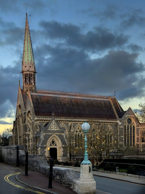 A vibrant bouquet arrangement of mixed fresh flowers, including pink roses, white lilies, and purple statice, is held by a person in front of a historic stone church with a tall, pointed spire, large