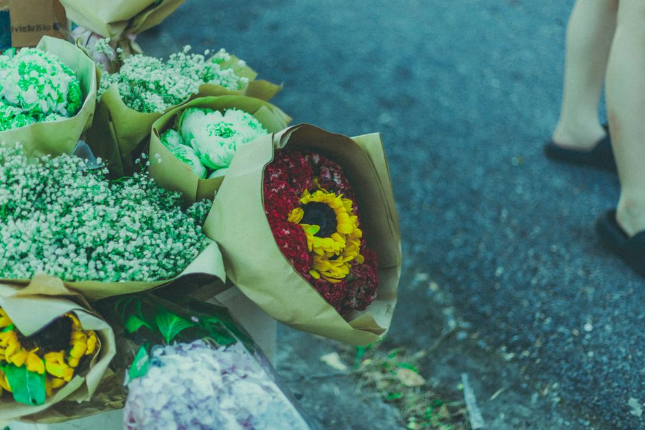 A bouquet arrangement of fresh flowers wrapped in beige paper is displayed on a dark outdoor surface. The bouquet features a variety of flowers including vibrant red and yellow blooms, white baby's br
