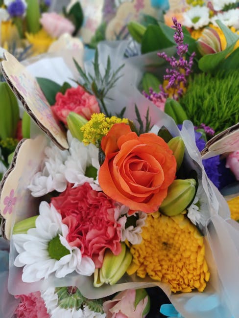 A vibrant floral bouquet featuring a variety of fresh flowers including an orange rose, yellow chrysanthemum, pink carnations, white daisies, and purple heather, arranged with greenery and wrapped in